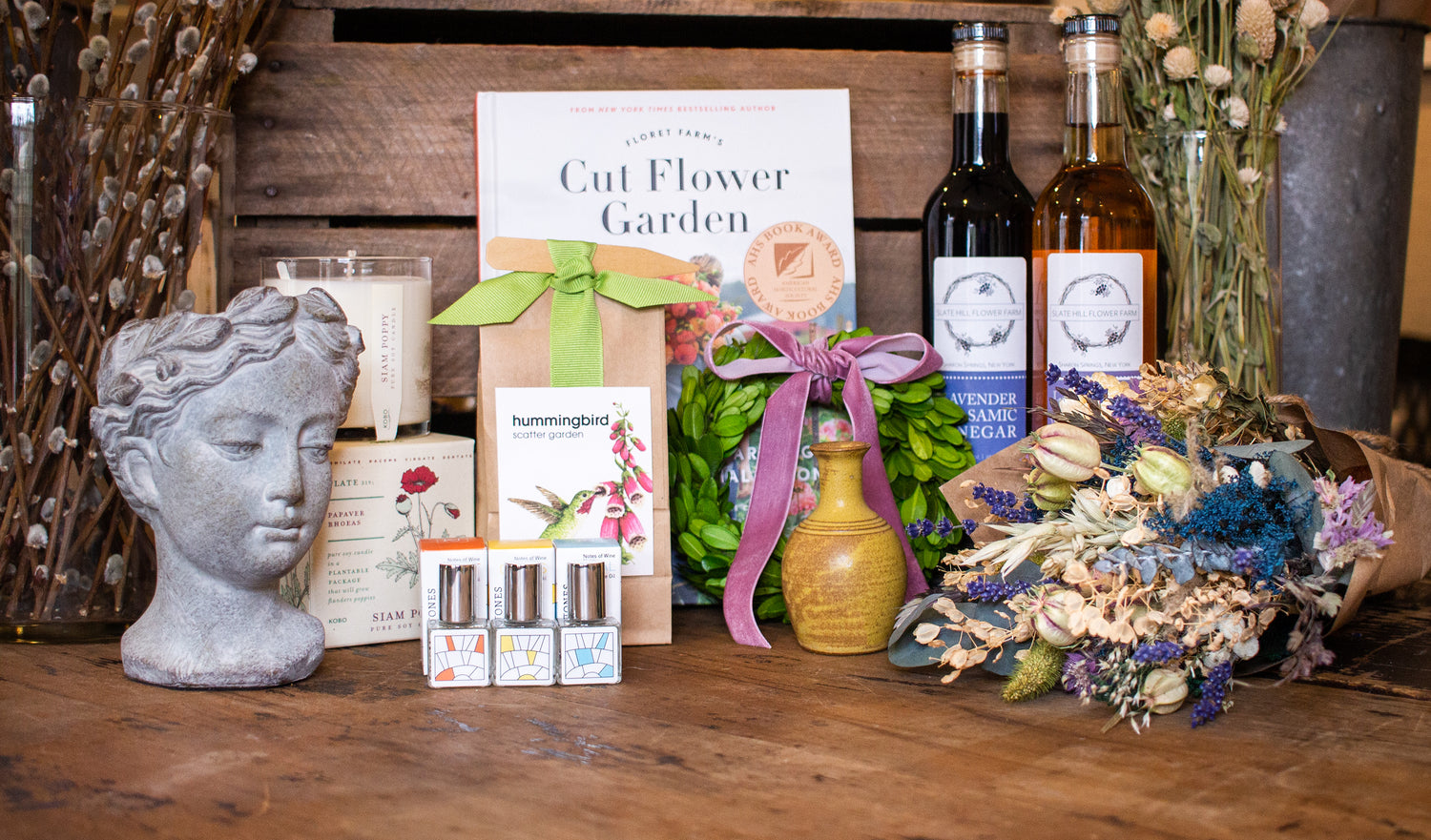 A display of various giftable items such as perfume, vases, seed packets, a dried flower bouquet, lavender balsamic vinegar, a "Cut Flower Garden" book, and candles.