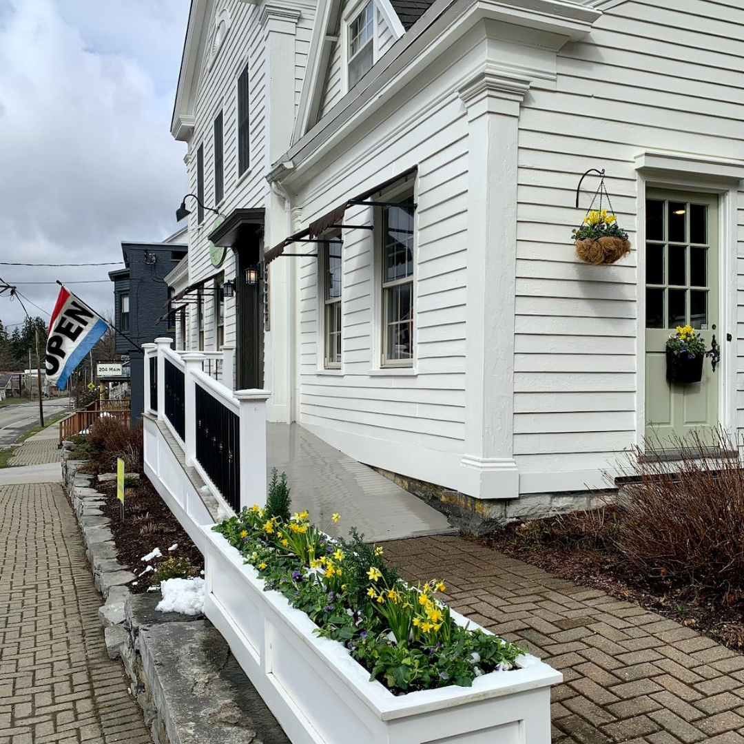 A side shot of the front of the building of Sharon Sprigs Fine Dried Flowers & Gifts retail shop. There are daffodils in a potted bed, a red white and blue OPEN flag waving in the wind, and the building is entirely white. The side door is a sage green while the front one is black. The railing is also mostly white but with black accents.