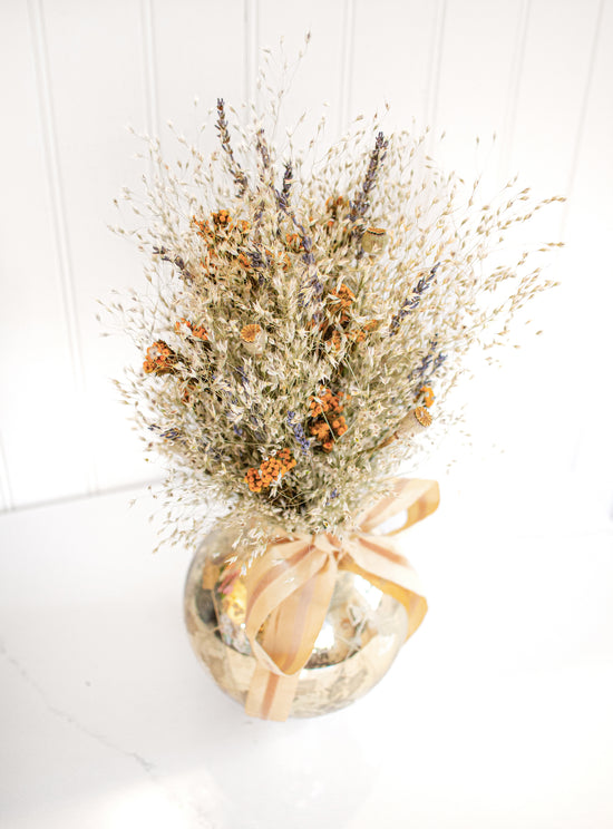 A custom dried flower piece. There is orange yarrow, wheat, and lavender inside a circular golden vase that has a light orange/tan bow tied at the mouth.