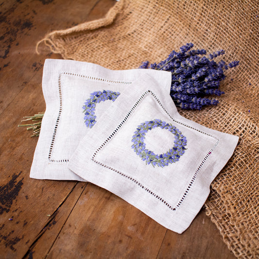 Two square, flat lavender sachets with a lavender ring embroidery that is sitting next to a lavender bundle, on top of a wooden table with burlap.