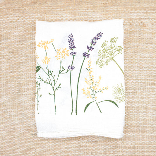 A folded June & December branded white tea towel with herb and flower designs.