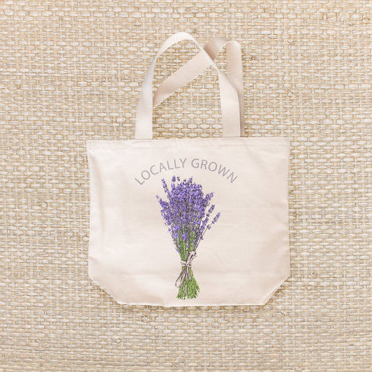 A natural colored canvas tote bag with text that reads "LOCALLY GROWN" and a graphic of a lavender bunch tied with string.