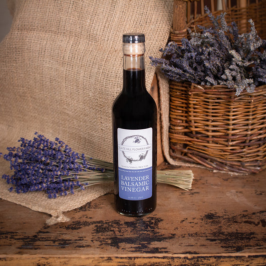 A glass bottle of Lavender Balsamic Vinegar and a label that reads "Slate Hill Flower Farm, Lavender Balsamic Vinegar". The bottle is on a rustic wooden table with a bunch of lavender sitting on a burlap piece on the left, as well as some lavender bunches in a wicker basket on the right.