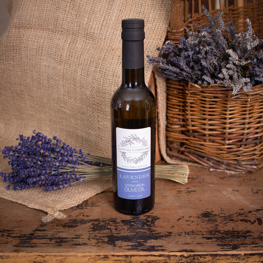 A glass bottle of Lavender Olive Oil and a label that reads "Slate Hill Flower Farm, Lavender Infused Extra Virgin Olive Oil". The bottle is on a rustic wooden table with a bunch of lavender sitting on a burlap piece on the left, as well as some lavender bunches in a wicker basket on the right.