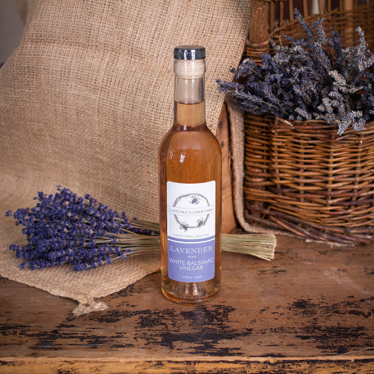 A glass bottle of White Balsamic Vinegar with a pink tint and a label that reads "Slate Hill Flower Farm, Lavender Infused White Balsamic Vinegar". The bottle is on a rustic wooden table with a bunch of lavender sitting on a burlap piece on the left, as well as some lavender bunches in a wicker basket on the right.