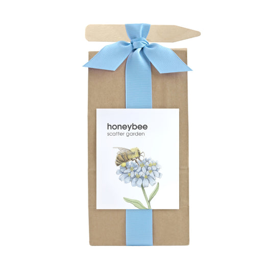 A long brown bag filled with scatter garden seeds. Wrapped with a light blue ribbon that also holds a garden marking stick. Label on the front has a bee & flowers graphic with text that reads "Honeybee Scatter Garden".