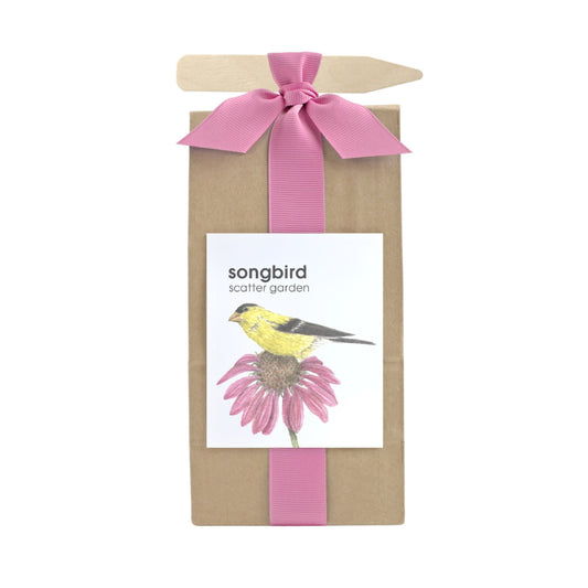 A long brown bag filled with scatter garden seeds. Wrapped with a pink ribbon that also holds a garden marking stick. Label on the front has a bird on top of a pink flower graphic with text that reads "Songbird Scatter Garden".