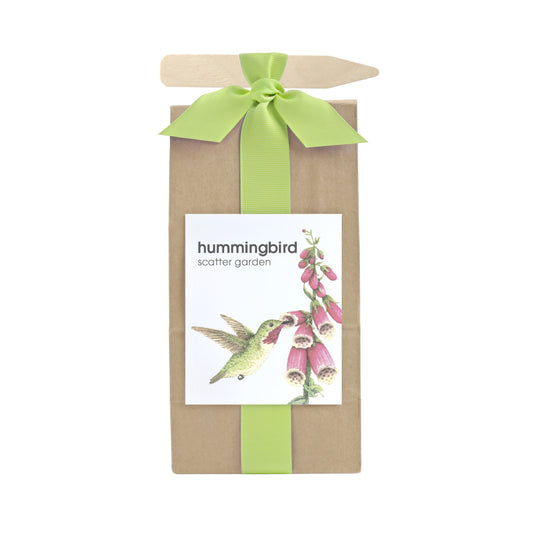 A long brown bag filled with scatter garden seeds. Wrapped with a green ribbon that also holds a garden marking stick. Label on the front has a hummingbird & flowers graphic with text that reads "Hummingbird Scatter Garden".