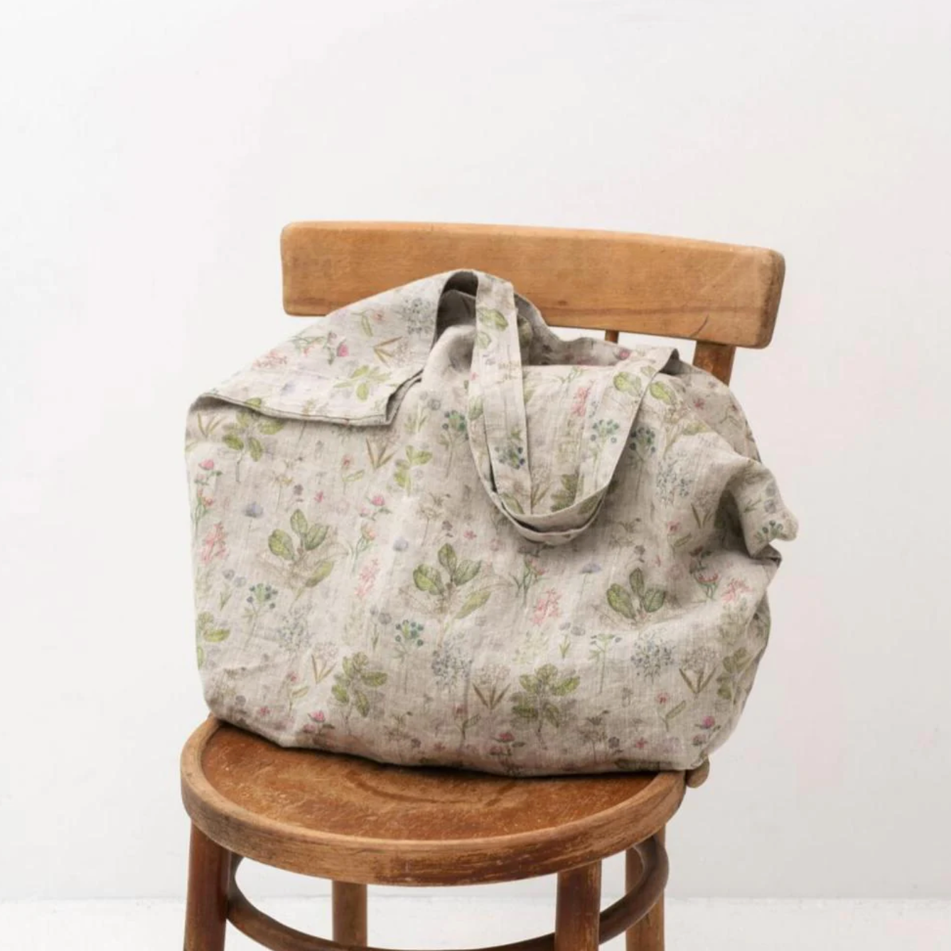 A Linen Tales Floral Linen Bag with pastel and green floral/leaf designs, sitting on a wooden chair,